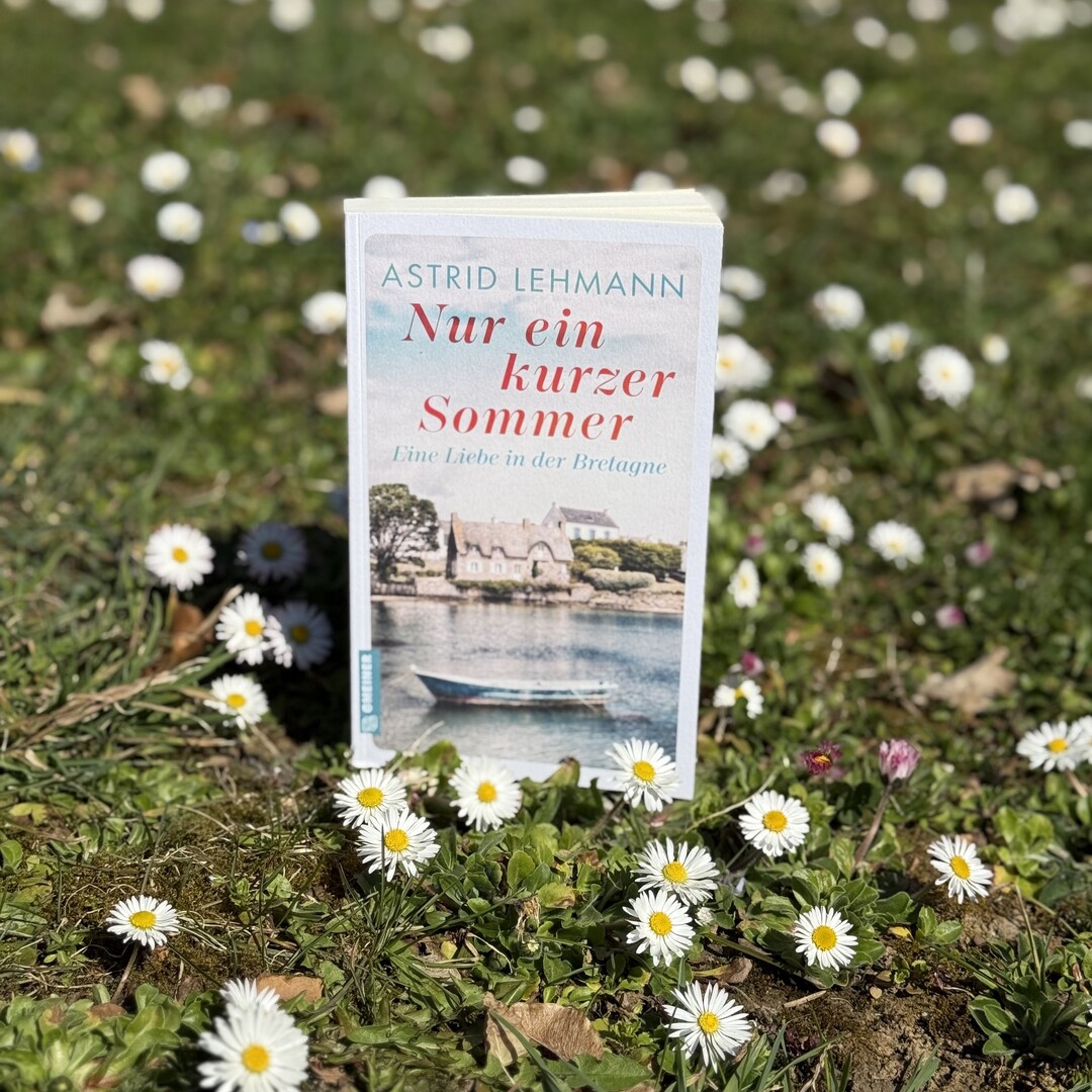 Auf einer grünen Wiese mit Gänseblümchen steht  das Buch "Nur ein kurzer Sommer" von Astrid Lehmann. Das Cover des Buches zeigt ein Ruderboot auf einem See, im Hintergrund ist ein altes Stein-Haus zu sehen.