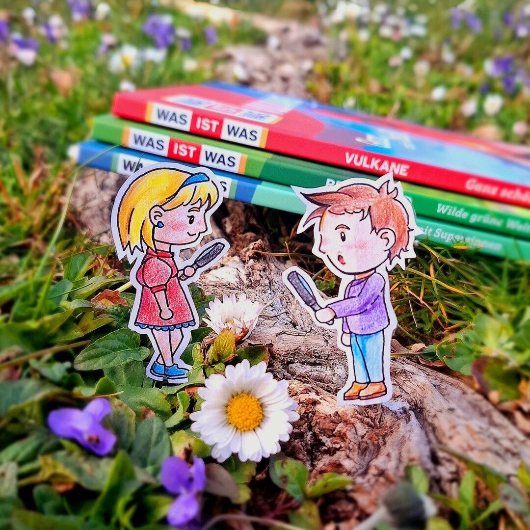 Drei "Was ist was"-Bücher liegen im gras, man sieht die Buchrücken. Davor stehen zwei Comic-Personen, die auf Papier gemalt und dann ausgeschnitten wurden. Sie halten Lupen in der Hand. Im Vordergrund ist ein Gänseblümchen zu sehen.