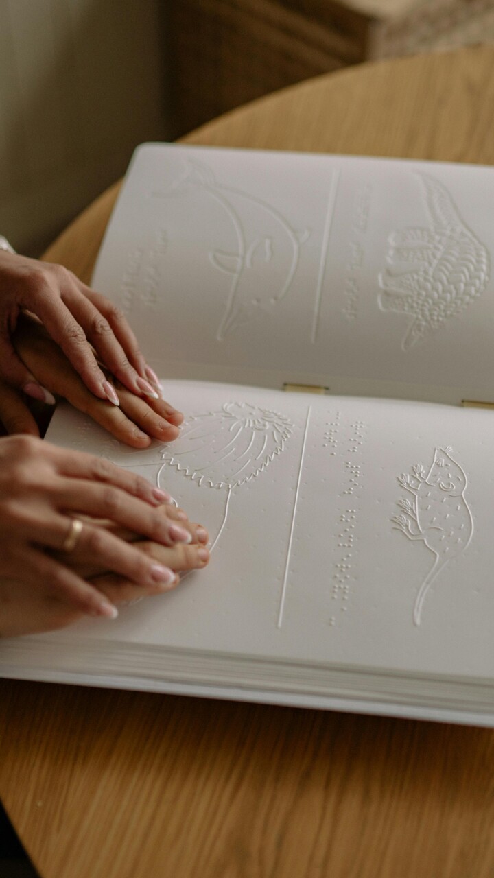 Ein Kind lässt seine Hände über ein Bilderbuch gleiten, dessen Motive geprägt sind. Geführt wird es dabei von den Händen einer erwachsenen Person.