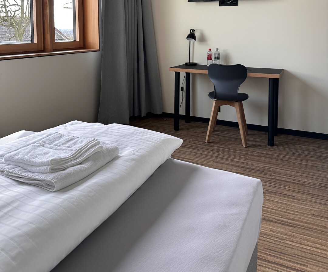 Zimmer am Mediacampus Frankfurt. Links befindet sich ein Fenster mit einem grauen Vorhang, der beiseite geschoben wurde. Das Bett hat weiße Bettwäsche und auf dem Bett liegen zwei weiße, gefaltete Handtücher. An der Wand gegenüber dem Bett steht ein Schreibtisch mit einer Schreibtischlampe und zwei Flaschen Wasser. Davor steht ein Stuhl.
