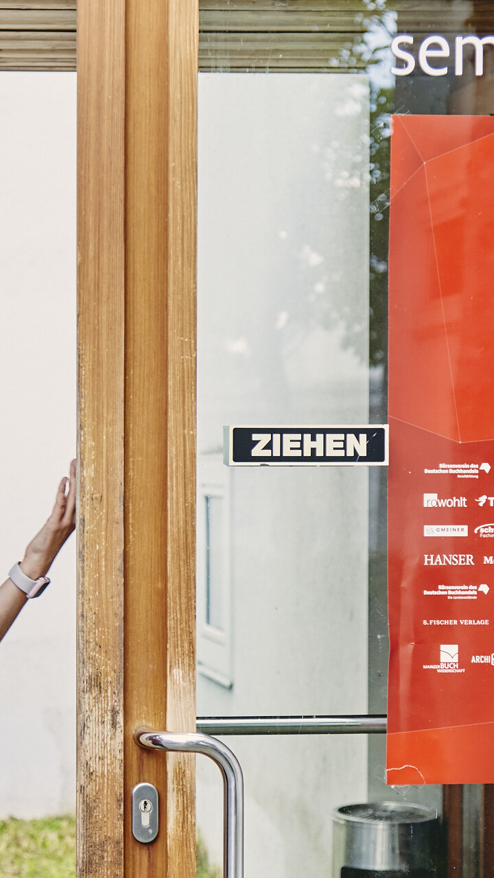 Zwei Schülerinnen betreten das Unterrichtsgebäude und lachen sich dabei an. Die Tür ist weit offen und an ihr hängt ein Plakat mit der Aufschrift "Junge Perspektiven der Buch- und Medienbranche".