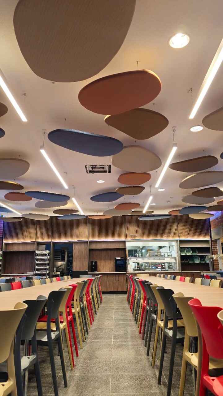 Mensa des Mediacampus Frankfurt. Zentraler Gang, links und rechts sind Stühle an Tische rangeschoben. An der Decke hängen Akoustik-Elemente in organischen Formen. Das Licht ist an und leuchtet die gesamte Mensa hell aus.